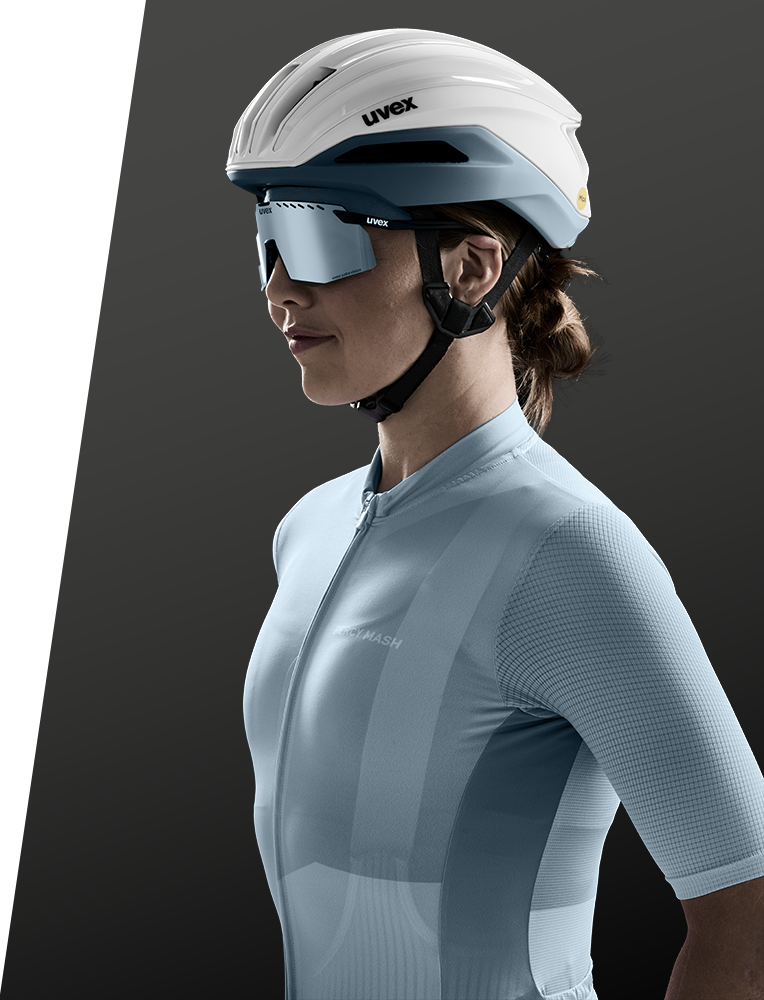 Eine Frau in hellblauer Radfahrerkleidung, weißem Helm und reflektierender Sonnenbrille posiert vor einem dunkelgrauen und weißen Hintergrund. Sie blickt nach links und ist von der Taille aufwärts fotografiert.