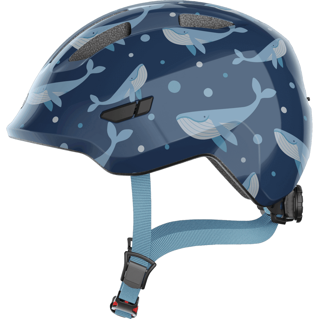 Der Abus Smiley 3.0 ist ein blauer Helm für Kleinkinder mit hellblauem und weißem Wal-, Blasen- und Wellenmotiv. Er verfügt über Belüftungslöcher und einen verstellbaren hellblauen Kinnriemen mit einer schwarz-roten Schnalle für zusätzlichen Schutz.